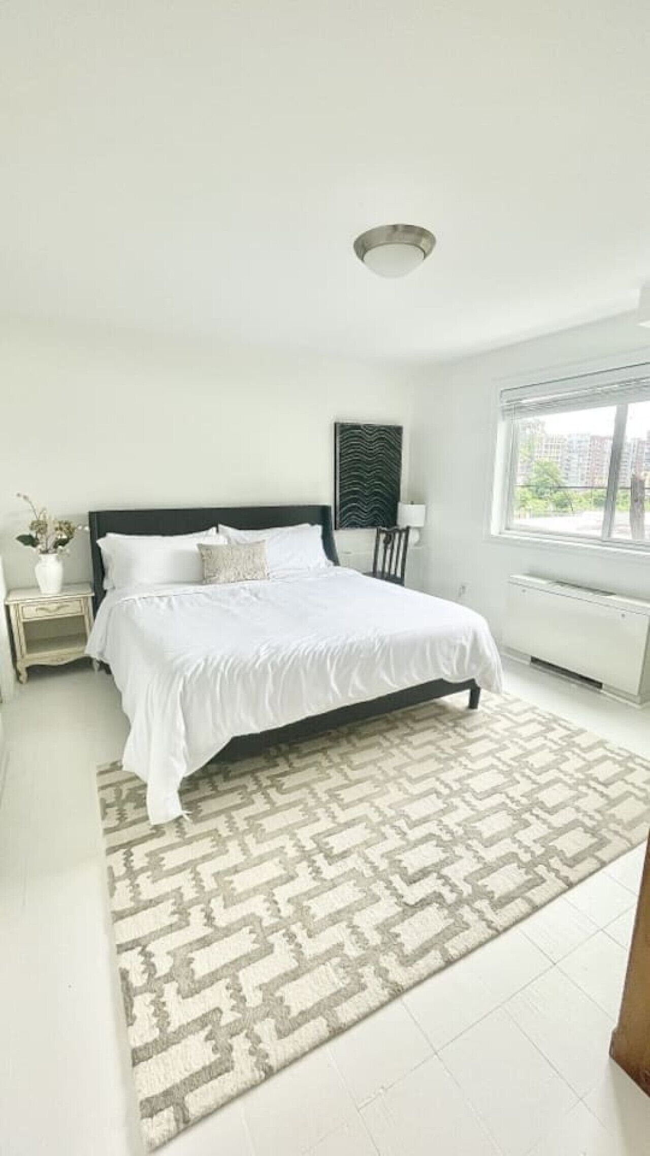 A bedroom with a bed, a nightstand, a radiator, a window, and a patterned rug.