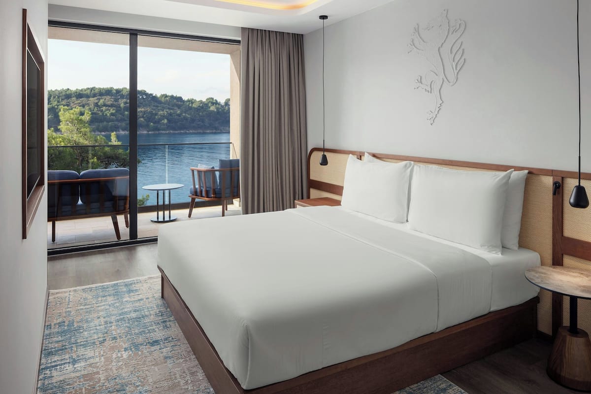 Une chambre d’hôtel moderne dotée d’un grand lit, d’un balcon avec coin salon et d’une vue sur un lac.