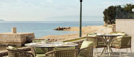 An outdoor dining area with wicker chairs and a round table, overlooking a beach and the sea.