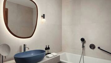 Un baño moderno con un espejo ovalado grande, un lavabo tipo recipiente azul, una bañera blanca y un cabezal de ducha de mano.