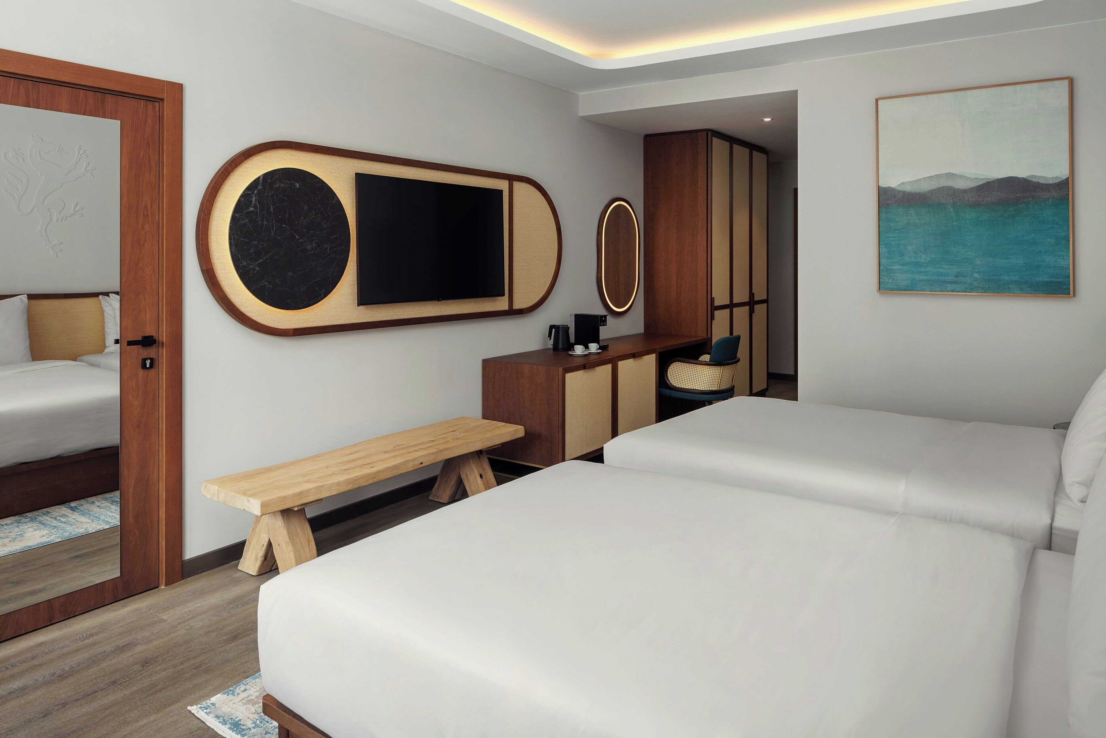A modern hotel room with a large bed, a bench, a desk with a coffee maker, and a painting on the wall.