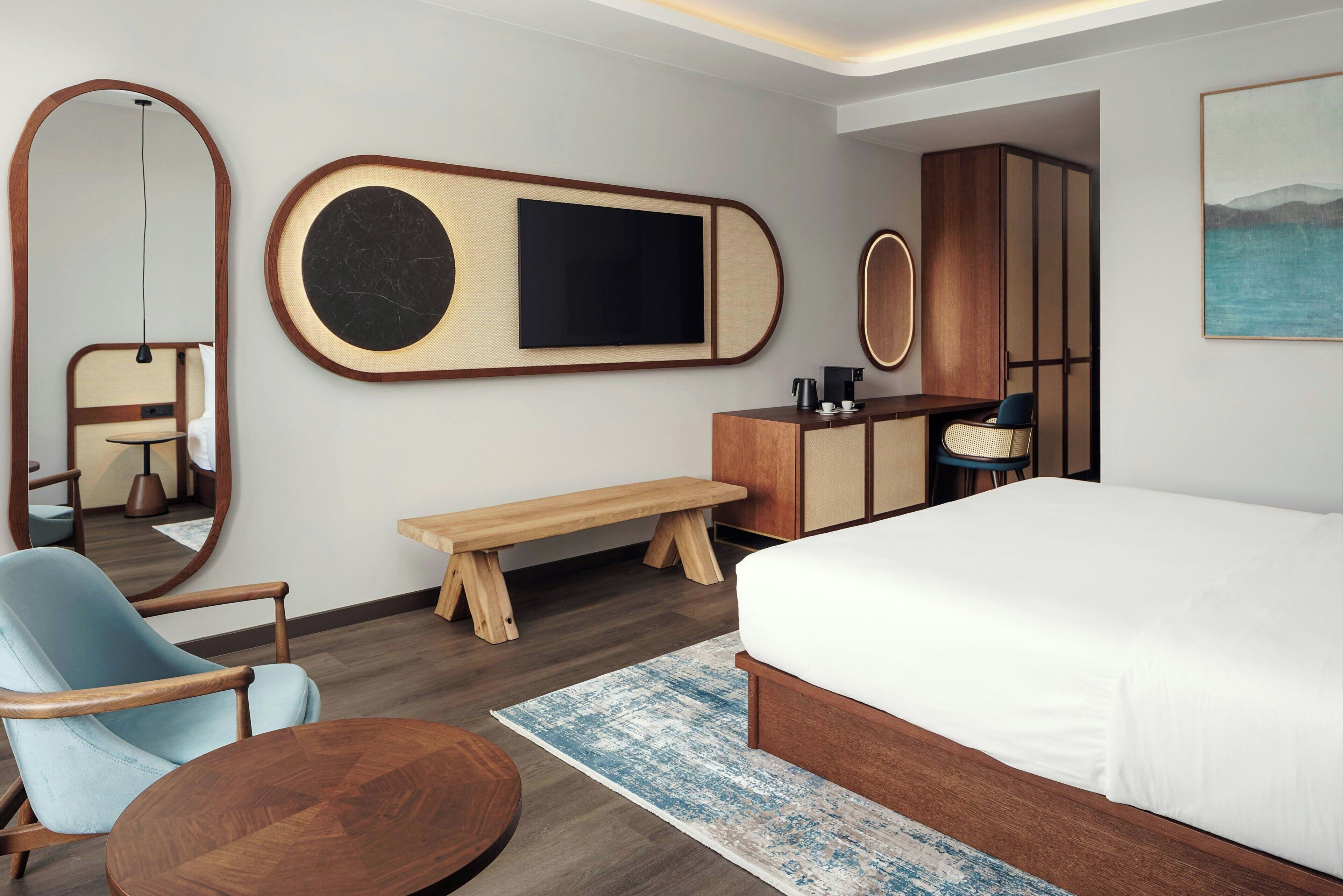 A modern hotel room with a large bed, a wooden bench, a round table, a chair, a TV, a desk, and a wardrobe.