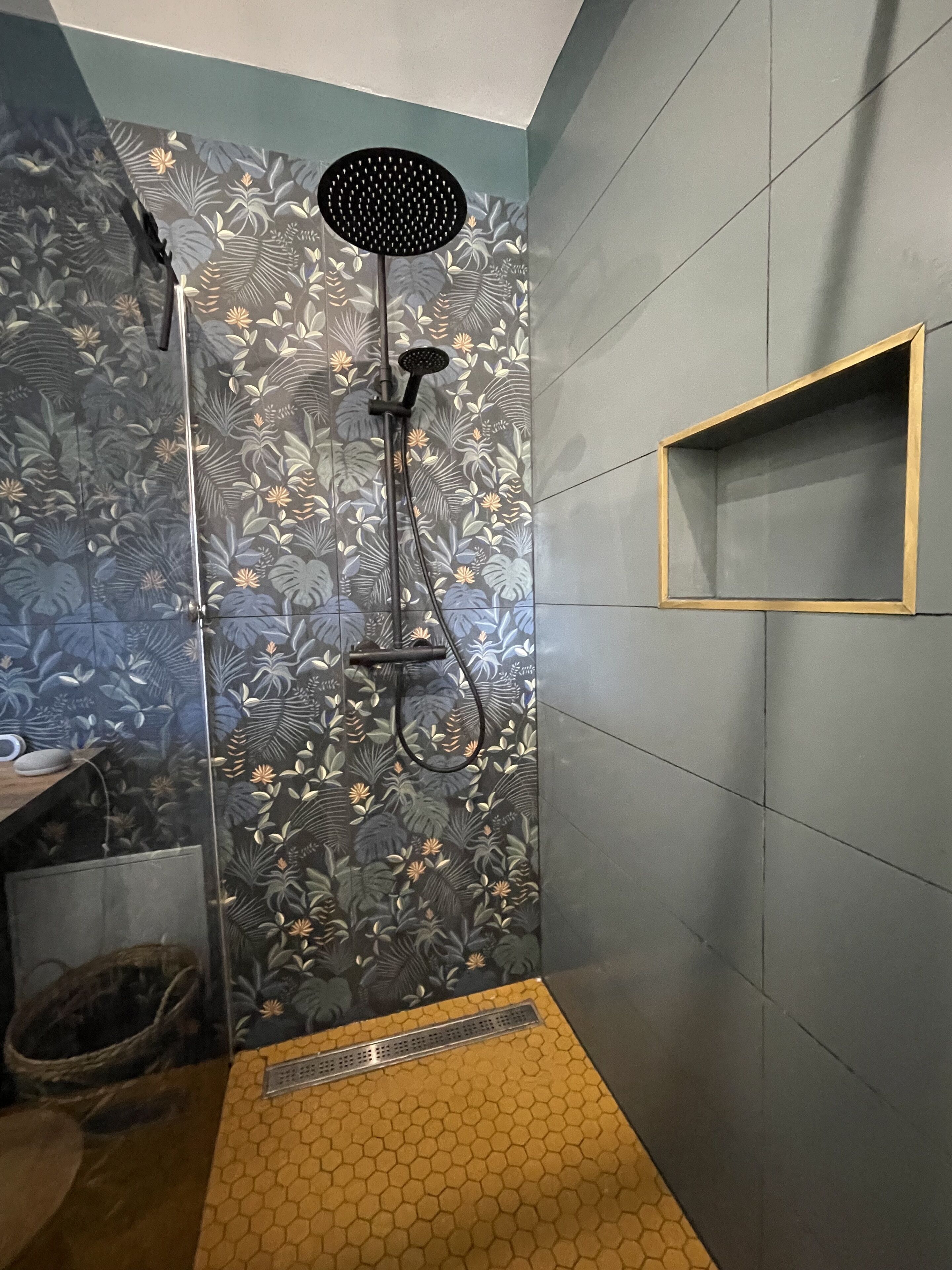 A shower with a floral tile pattern, a yellow hexagonal floor, and a small wooden shelf.