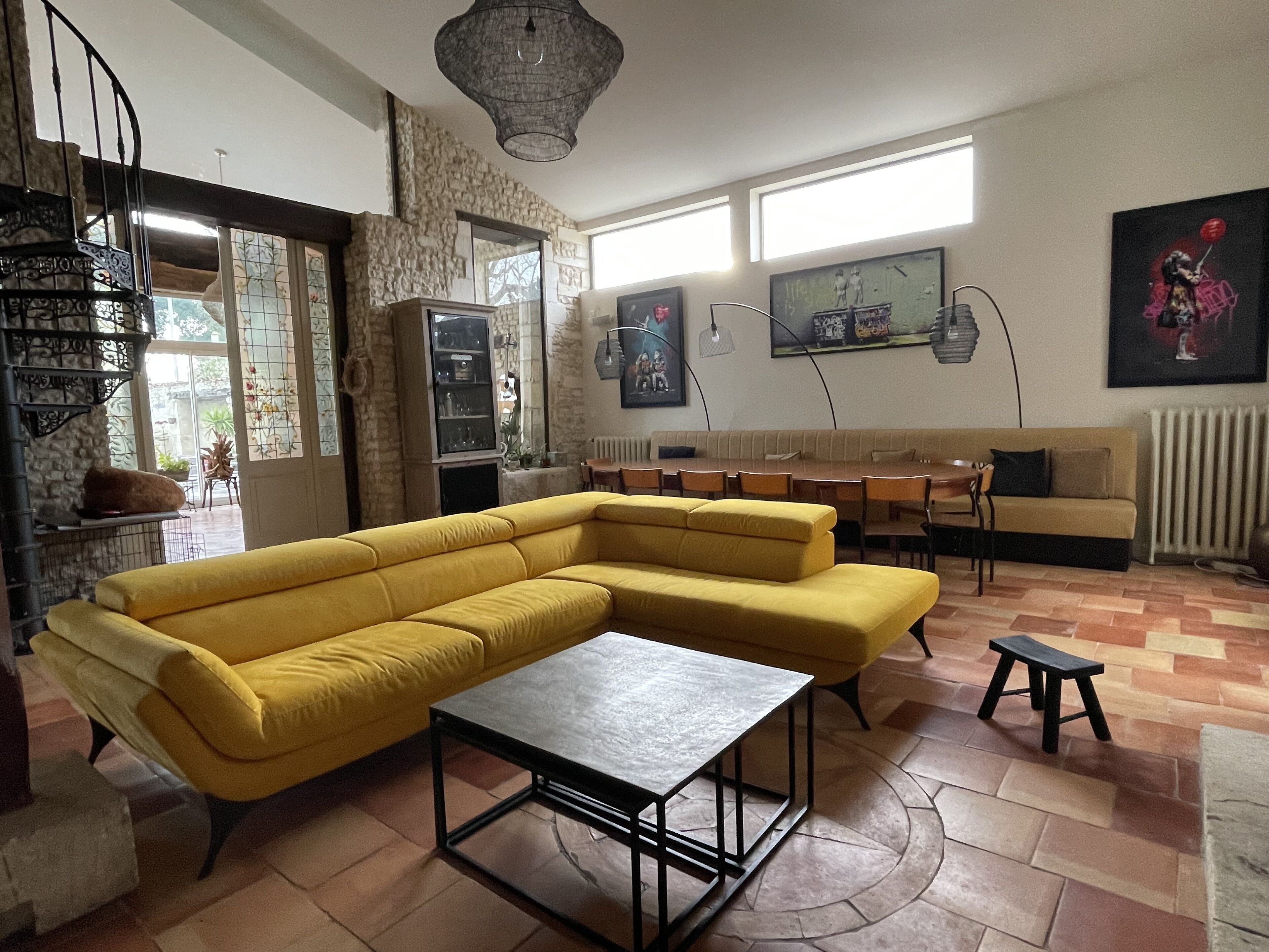 A living room with a yellow sectional sofa, a coffee table, a dining area with a table and chairs, a staircase, and a large window providing natural light.