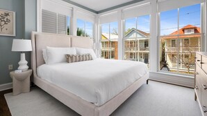 A spacious bedroom with a large bed, a nightstand, a dresser, and a view of the neighborhood through large windows. - 3 Luxury Houses w/ Patios | 1 Block from King St (Charleston)