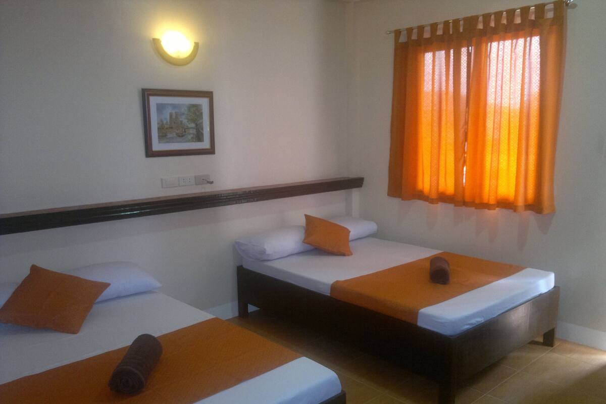Een hotelkamer met twee enkele bedden, elk met een oranje deken en kussen. Boven de bedden hangt een ingelijst schilderij, en er is een raam met oranje gordijnen.