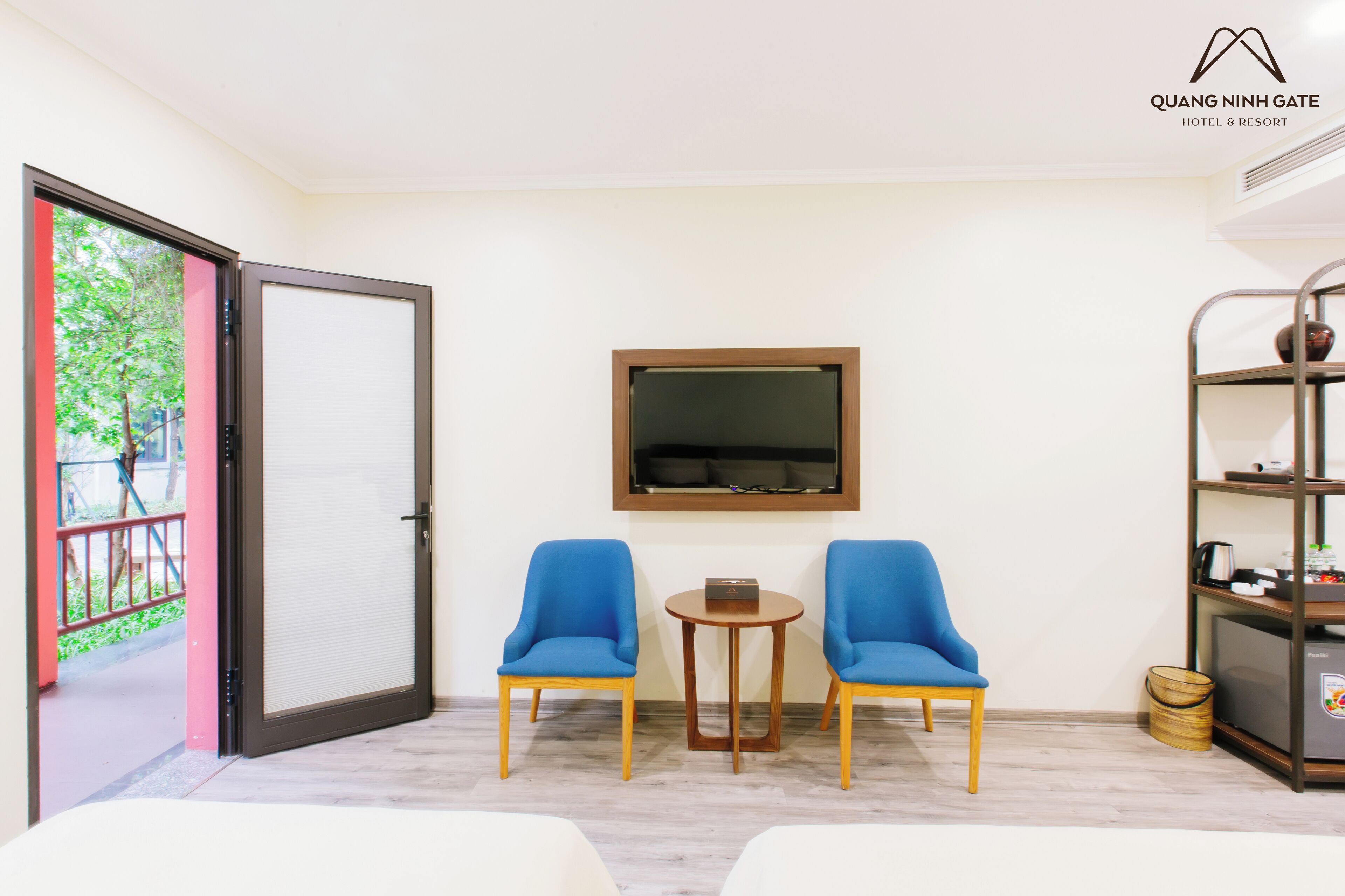 A hotel room with a sliding glass door leading to a balcony, two blue chairs around a small wooden table, a mounted television, and a bookshelf with various items.