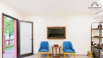 A hotel room with a sliding glass door leading to a balcony, two blue chairs around a small wooden table, a mounted television, and a bookshelf with various items.
