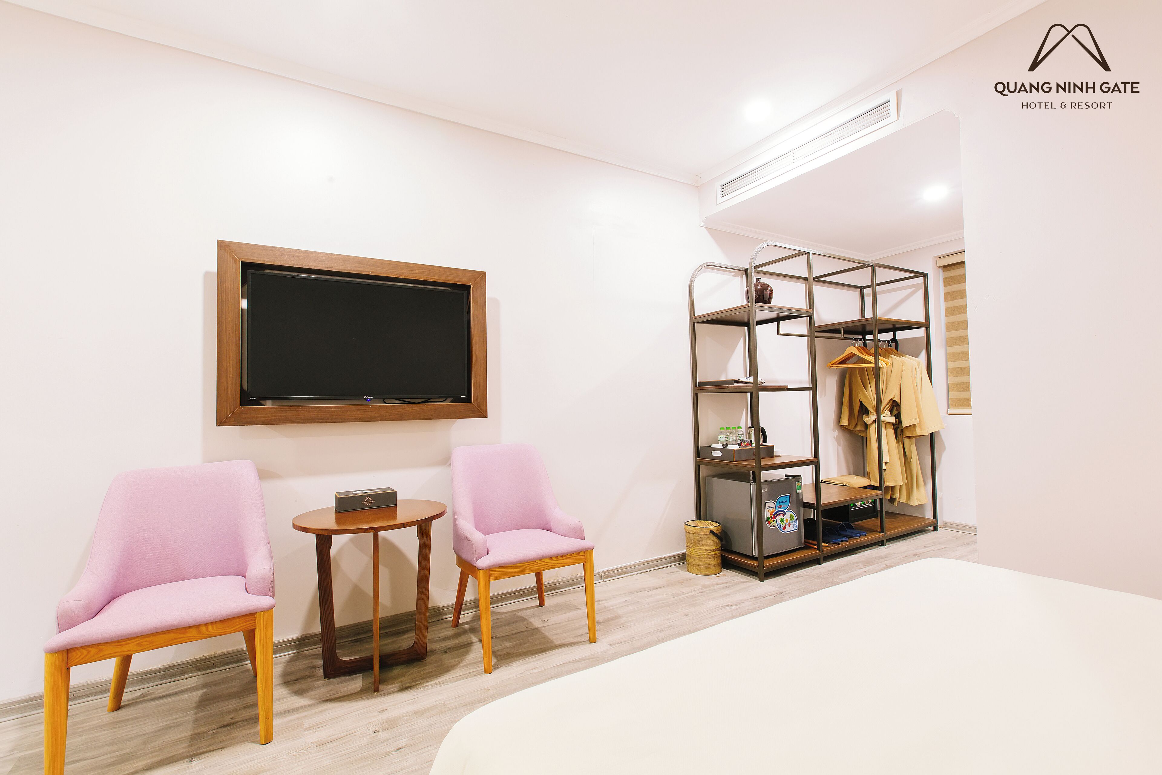 Una habitación de hotel moderna con una cama, dos sillones rosa, una mesita, un televisor, un perchero y un mueble de almacenamiento.