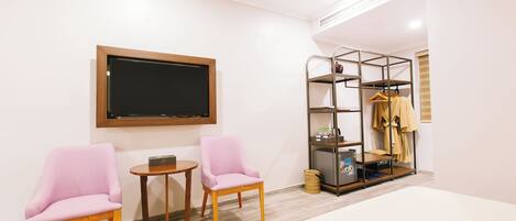 A modern hotel room with a bed, two pink armchairs, a small table, a TV, a coat rack, and a storage unit.