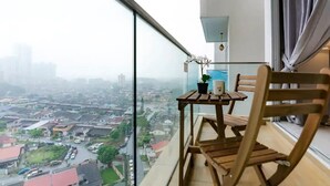 Een balkon met uitzicht op een stadsgezicht, een houten stoel, een klein tafeltje met een plant en een mok, en een raam met gordijnen.