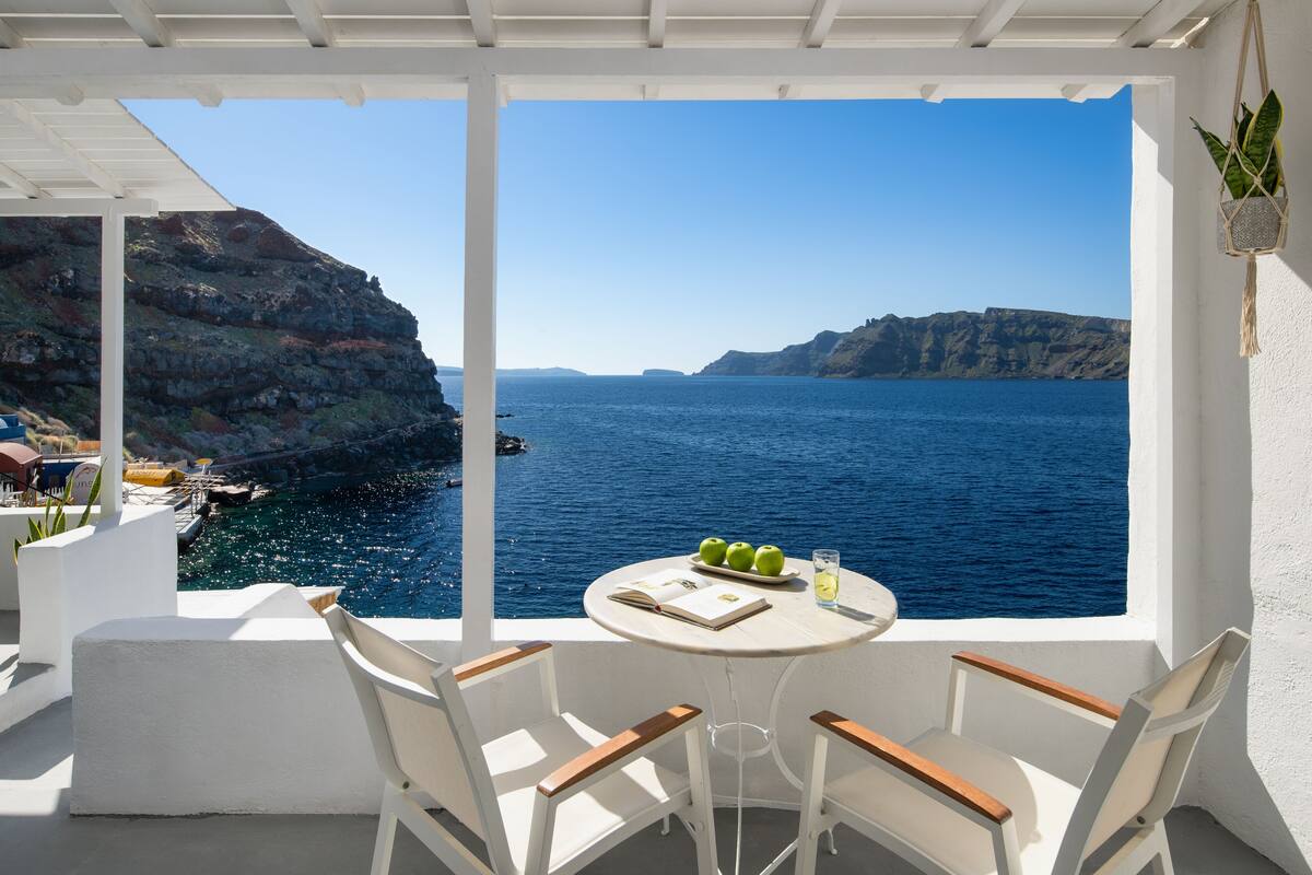 A terrace with a white table set for two, overlooking a blue sea and rocky cliffs.