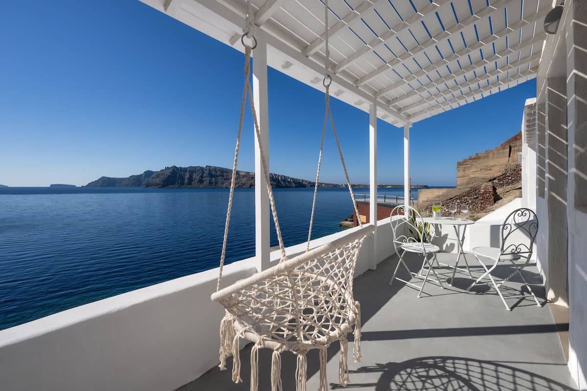A white outdoor seating area with a hanging chair and metal chairs, overlooking a body of water and rocky cliffs.