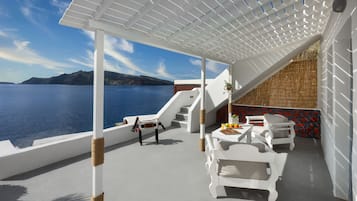Un'area salotto esterna riparata con mobili bianchi, un tavolo con frutta e vista sul mare.