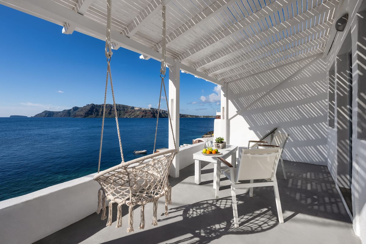 A balcony with a white wicker chair, a table with fruit, and a view of the sea and a distant island.