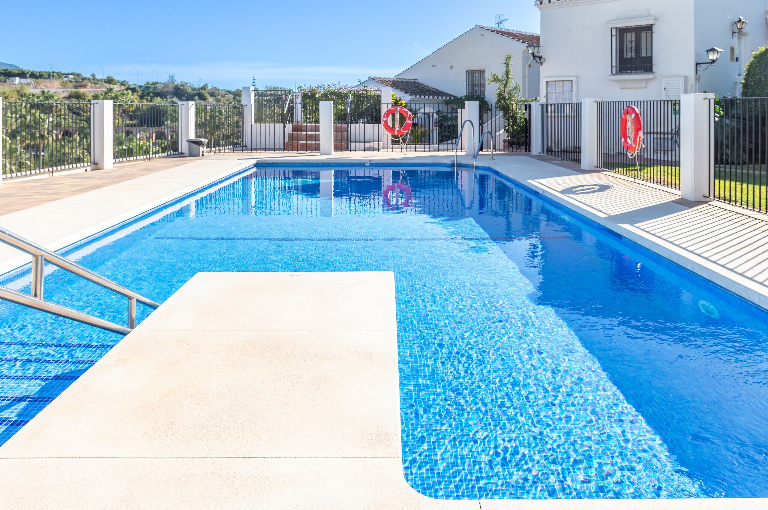 A swimming pool with a clear view of the surrounding area, including a white building with a tiled roof and a fenced garden.