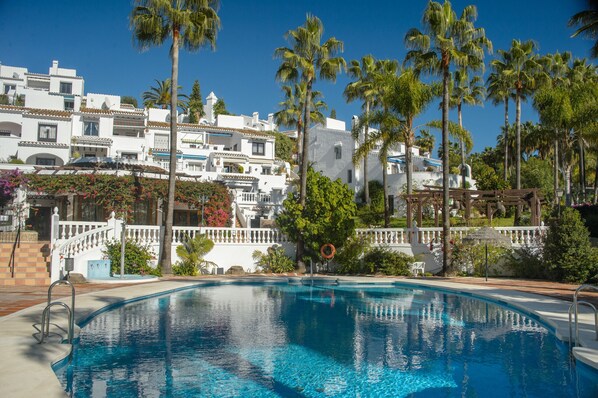 A swimming pool with a white fence, palm trees, and a building with white walls and red-tiled roofs.