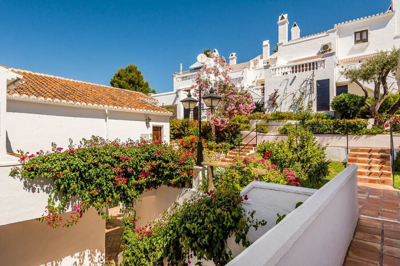Un edificio blanco con techo de tejas, una escalera y un jardín con flores rosas.
