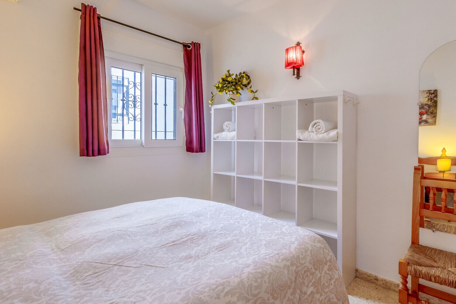 Un dormitorio con cama, armario, ventana con cortinas, lámpara de pared y una mesita con lámpara.