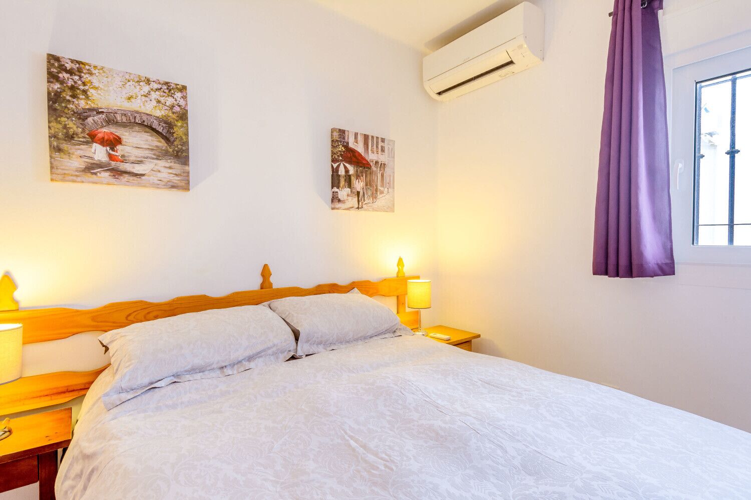 Camera d'albergo con un letto, due comodini, un tavolino, un condizionatore d'aria, una finestra con tende viola e due quadri appesi al muro.