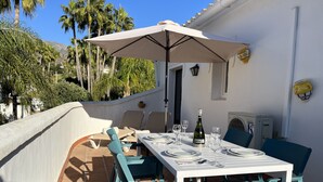 Terrassa a l'aire lliure amb taula preparada per dinar, una ampolla de xampany i un parasol gran.