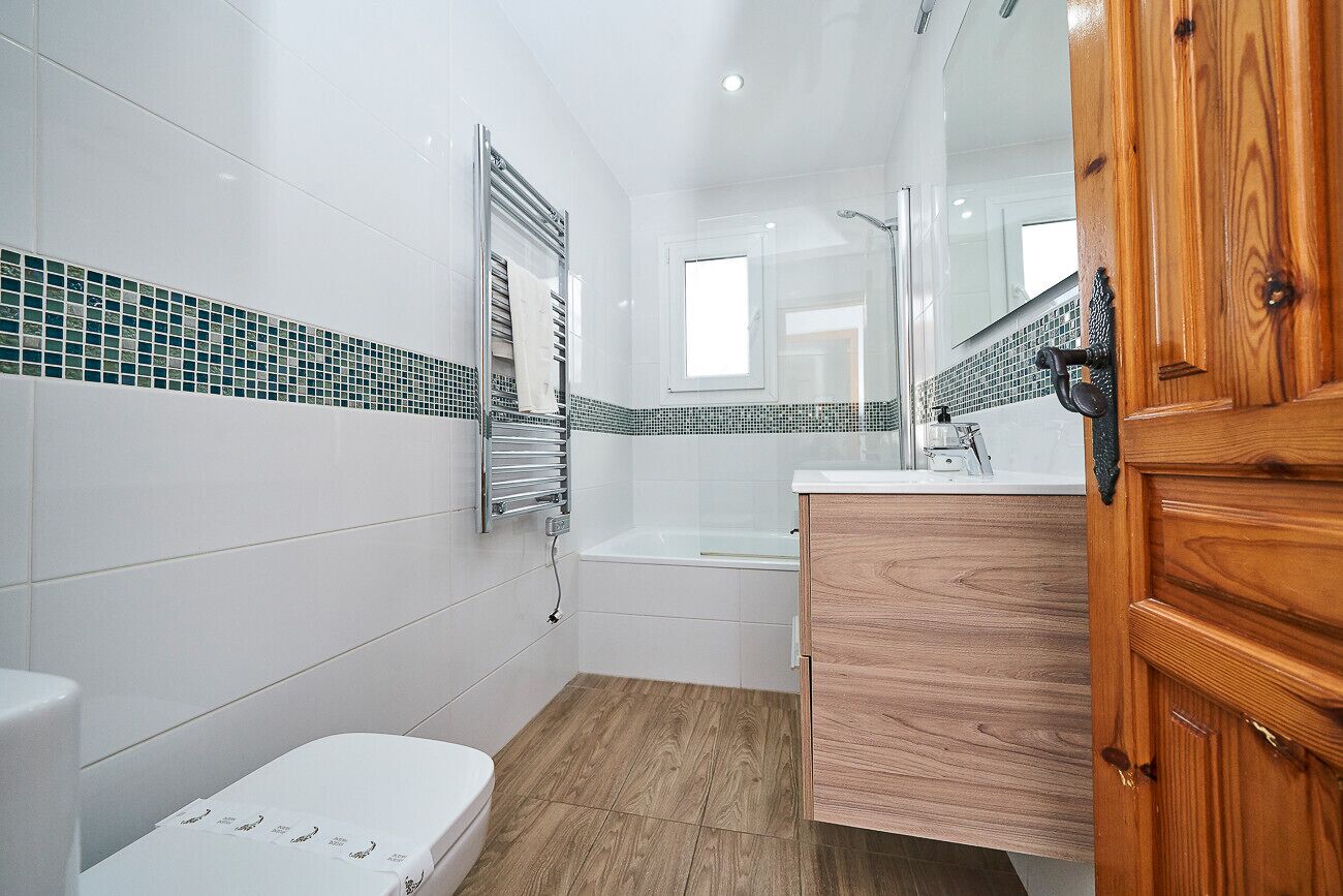 A modern bathroom with white tiles, a wooden vanity, a glass shower enclosure, and a visible toilet.