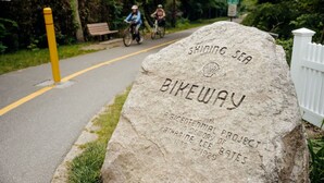 A stone marker with 'Shoddy Sea Bikeway' and 'Bicentennial Project' inscribed, surrounded by greenery and a pathway.