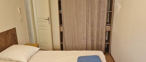 A neatly made bed with a folded blue towel, a wooden headboard, a white door, a small shelf, and a television mounted on the wall.