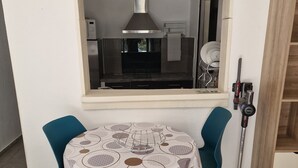 Un área de comedor con una mesa preparada para dos, una ventana que da a una cocina y una aspiradora en la esquina.