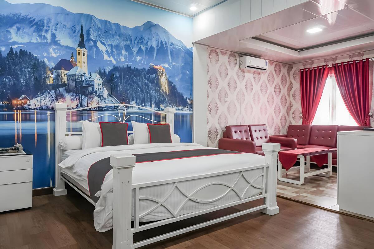 A bedroom with a bed, a red sofa, a small table, and a wall mural of a castle by a lake.