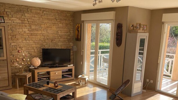 A living room with a stone wall, a flat-screen TV, a sofa, a coffee table, and a balcony with a chair. - SEVRIER, Bel appartement de 73M2 de 2 chambres pour 4 personnes avec parking et terrasse avec vue sur les montagnes, situé à 500m de la piste cyclable et du lac... (Sévrier)