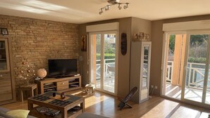 A living room with a stone wall, a flat-screen TV, a wooden coffee table, a sofa, and a balcony with a railing. - SEVRIER, Bel appartement de 73M2 de 2 chambres pour 4 personnes avec parking et terrasse avec vue sur les montagnes, situé à 500m de la piste cyclable et du lac... (Sévrier)