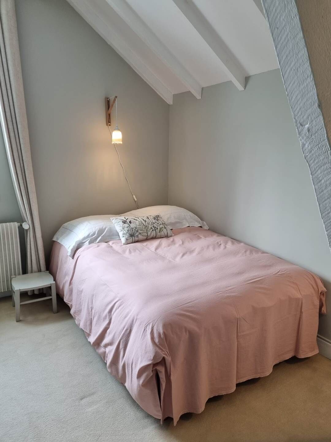 Een slaapkamer met een schuine balklaag, een bed met roze beddengoed, een nachtkastje en een wandlamp.