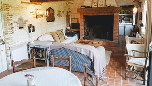 Ein rustikaler Raum mit freiliegenden Backsteinwänden, einem Kamin, einem Bett, einem Esstisch und einem durch eine offene Tür einsehbaren Küchenbereich.