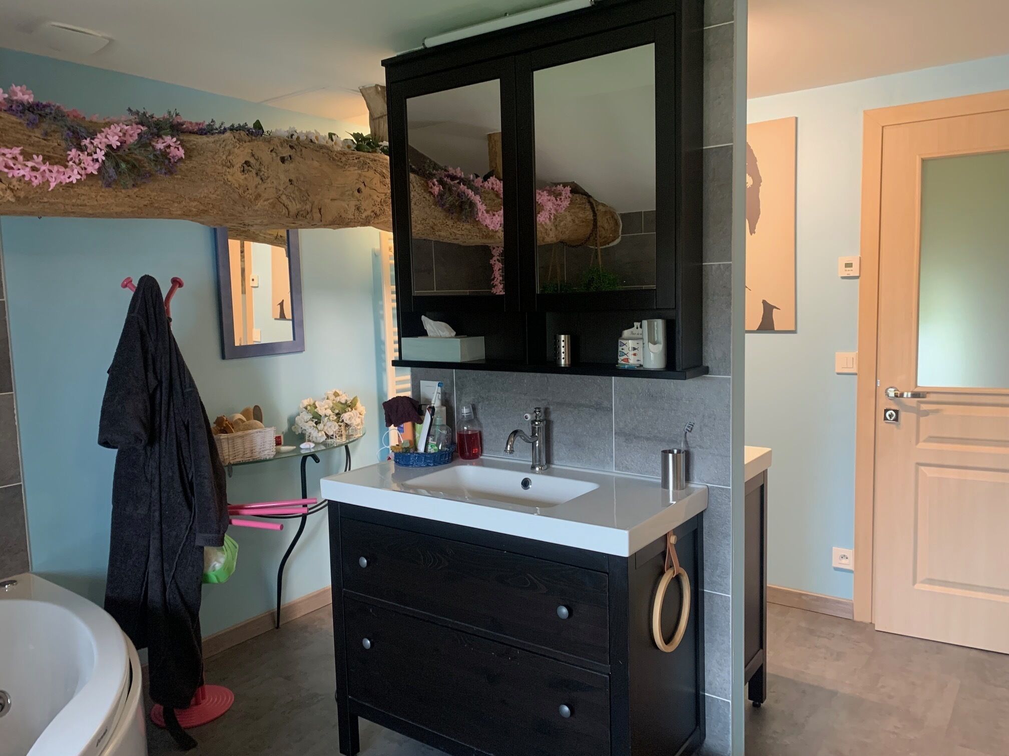 A bathroom with a white sink, black cabinet, and a mirror above it. There is a towel rack with a towel hanging on it, and a small shelf with toiletries.