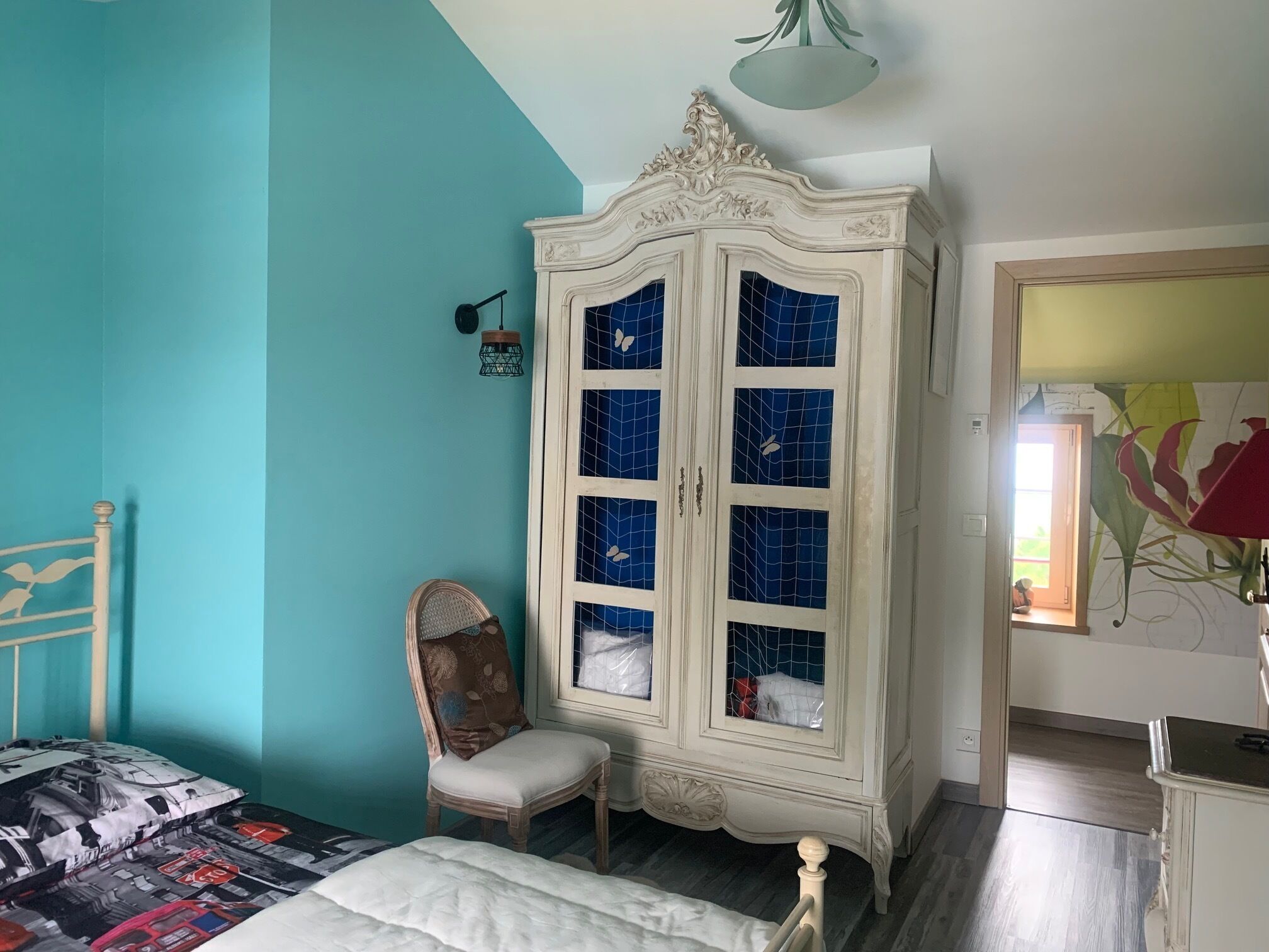 A bedroom with teal walls, a white wardrobe with blue-tiled doors, a wooden bed, and a chair.