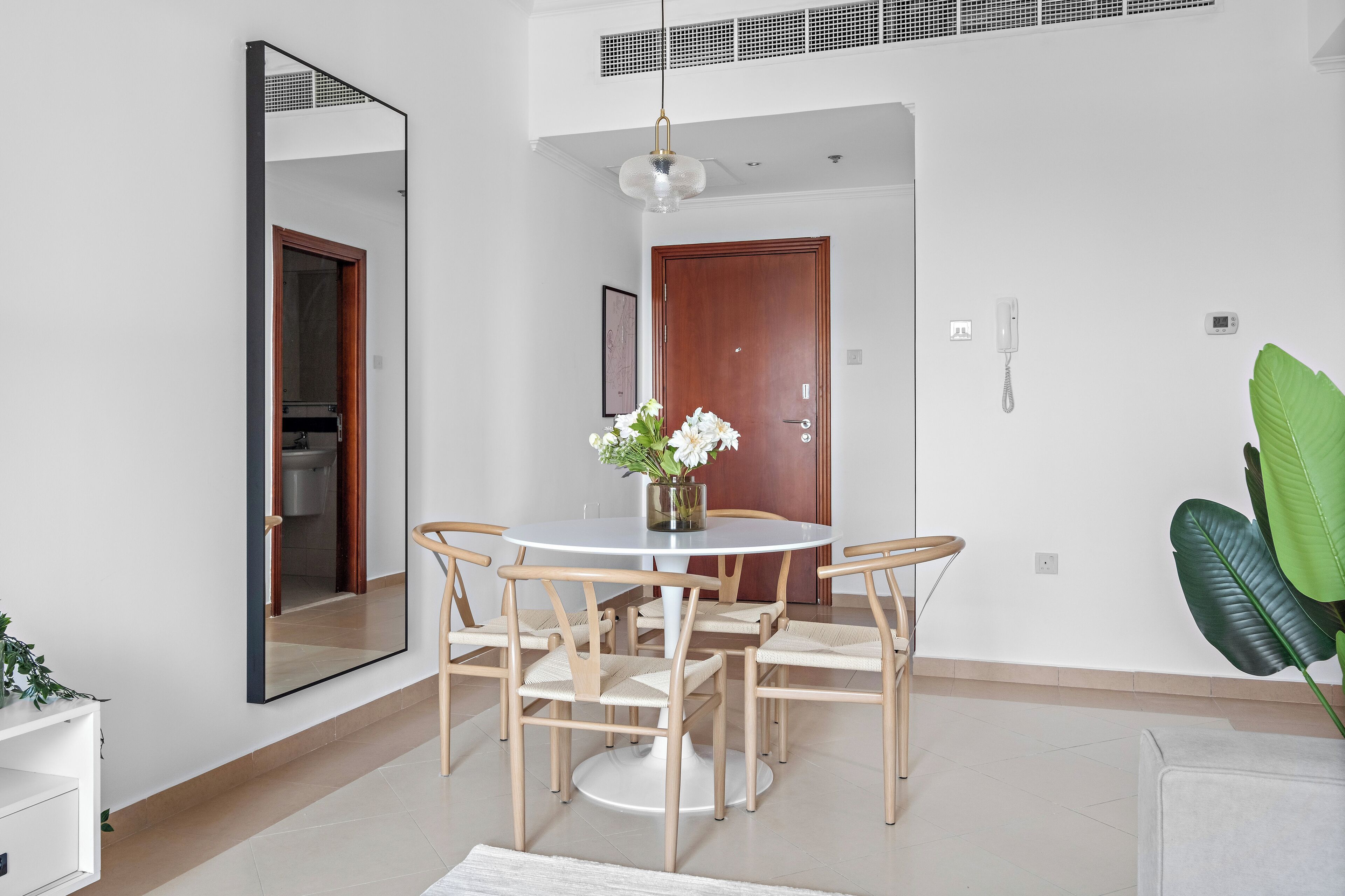 A modern dining area with a round table, six chairs, a vase with flowers, a large mirror, a wooden door, and a green plant.