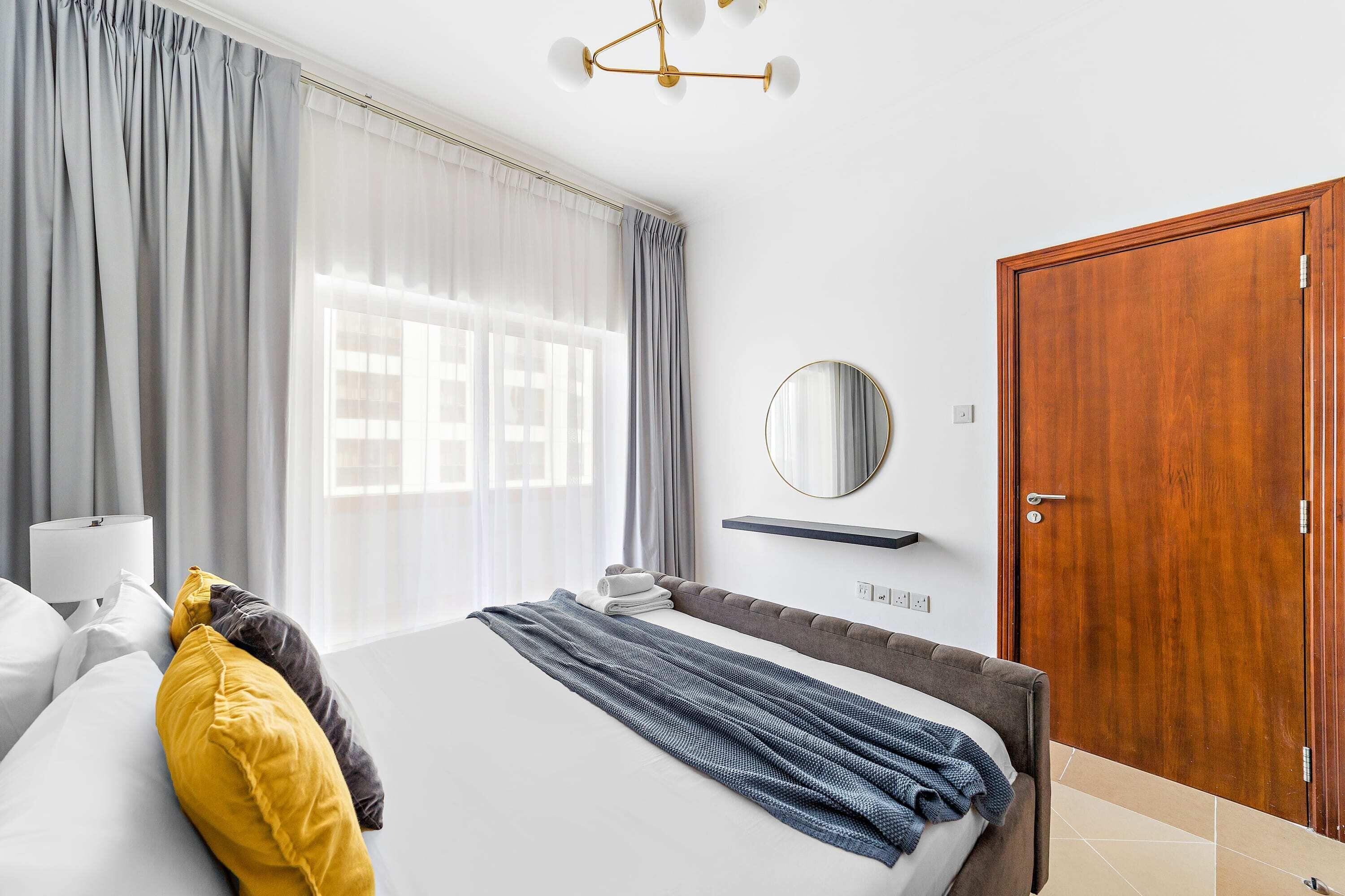 A hotel room with a bed, curtains, a bedside table, a lamp, a mirror, and a wooden door.