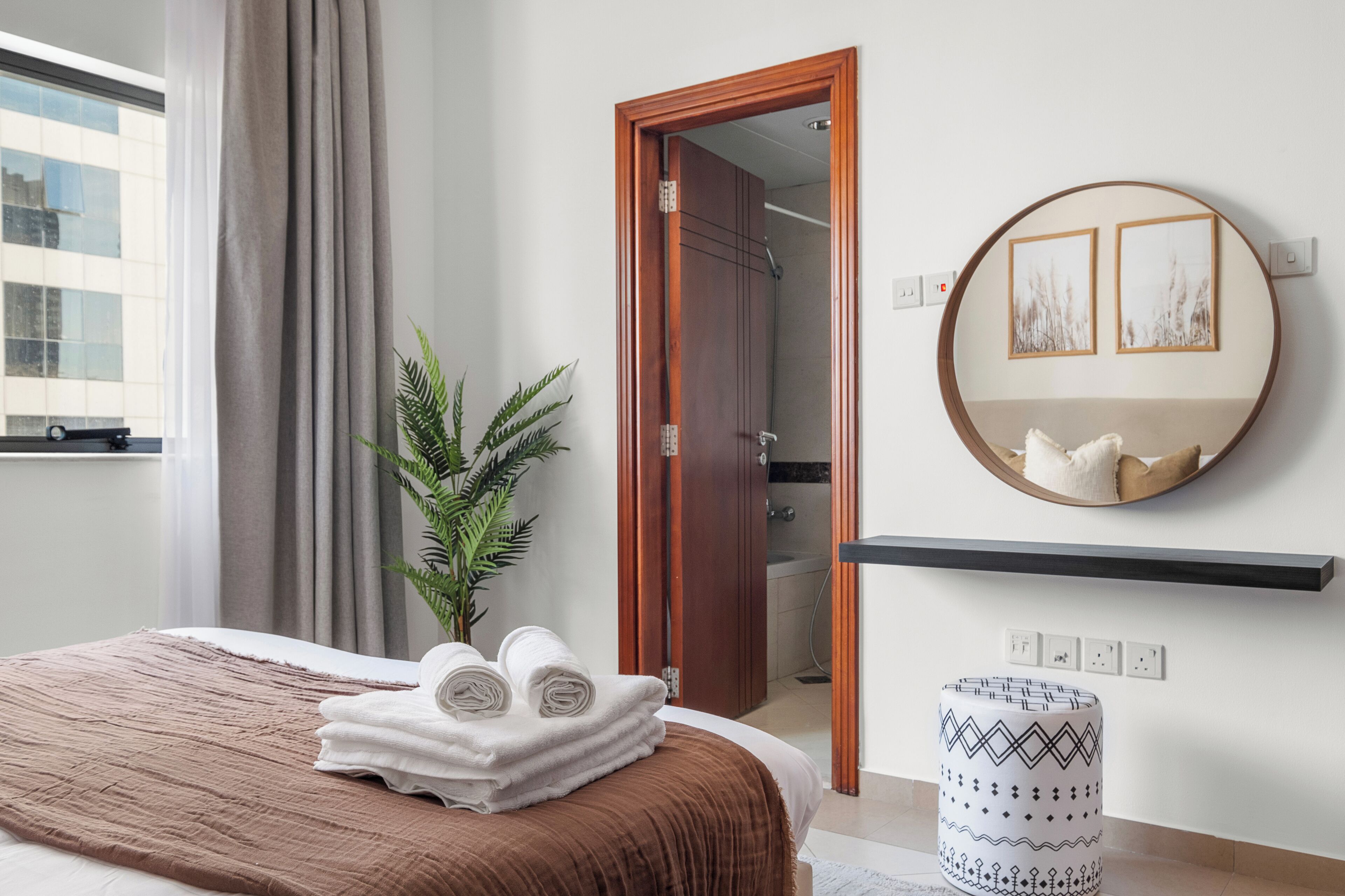 A hotel room with a bed, a mirror, a plant, and a view of the outside.