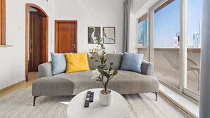 Ein modernes Wohnzimmer mit einem grauen Sofa, einem weißen Couchtisch und einer Topfpflanze. Ein großes Fenster bietet Blick auf Gebäude.