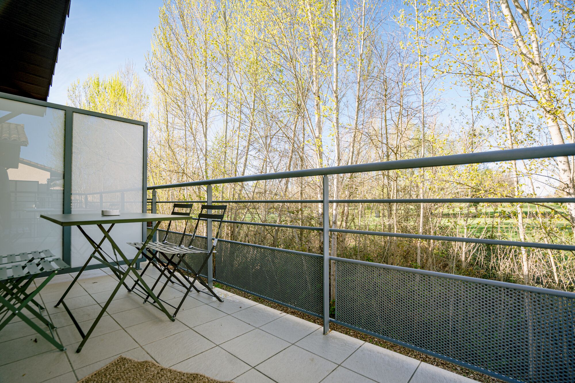 A balcony with a table and chairs, a metal railing, and a view of trees and a green field.
