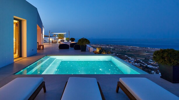 A modern outdoor pool area with a view of the ocean at dusk, featuring a seating area with lounge chairs and a small table.