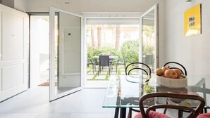 Uma área de refeições moderna com mesa e cadeiras de vidro, porta branca que dá para um jardim e quadro amarelado na parede.