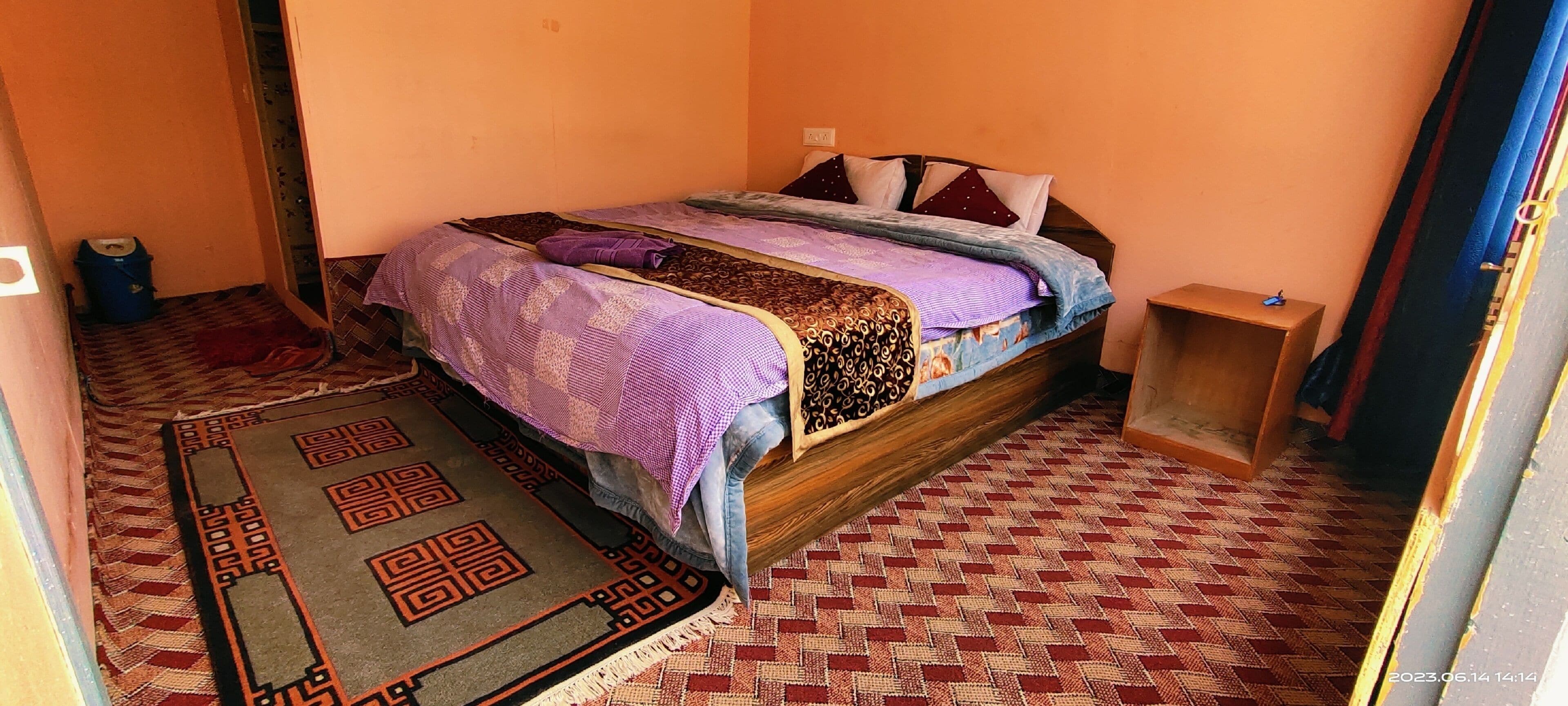A single bed with a patterned bedspread, a small bedside table, and a rug on a patterned floor.