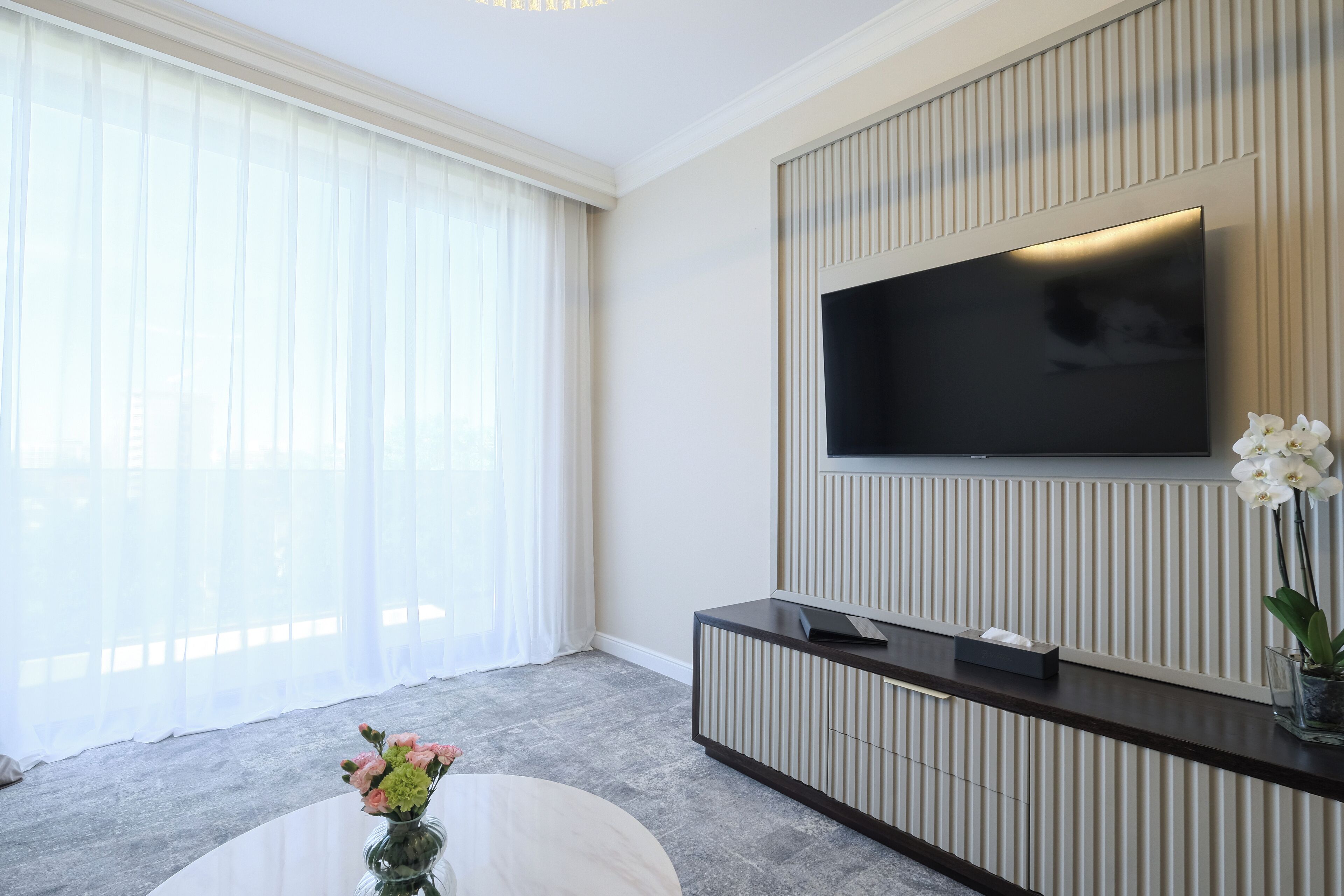 A modern living room with a large flat-screen TV mounted on a wall, a dark wooden console table, and a vase with flowers on a round coffee table.