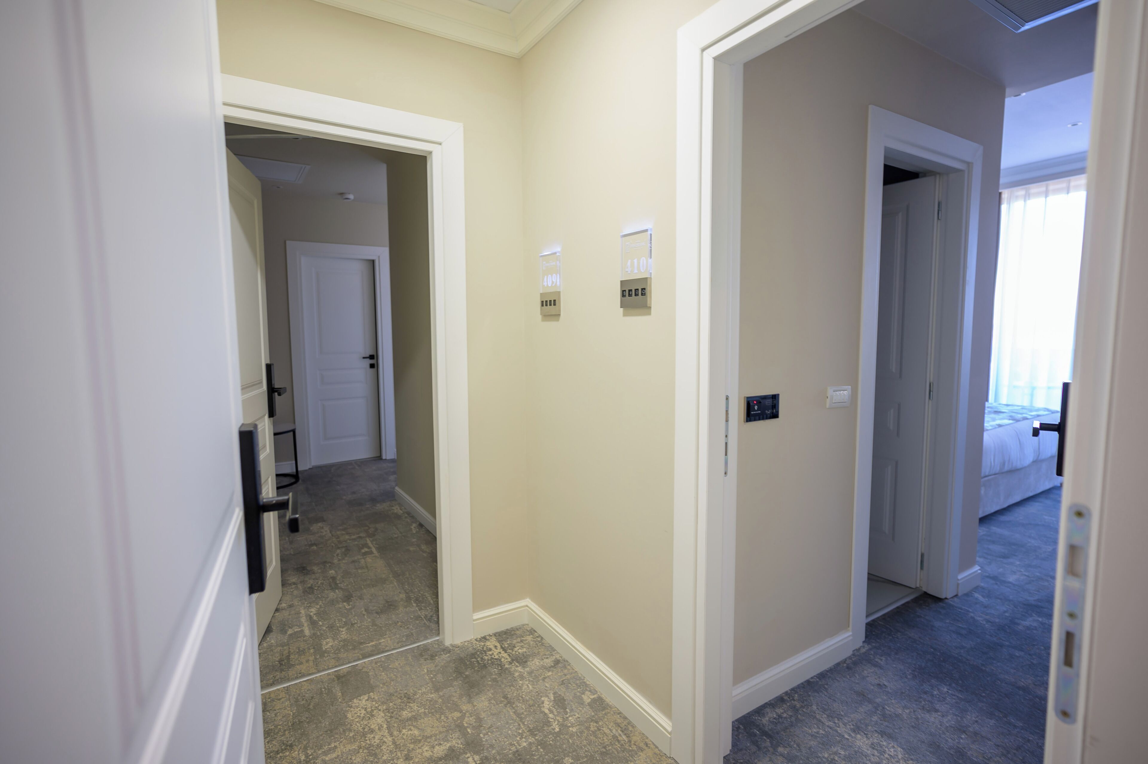 Un couloir aux murs de couleur beige, aux boiseries blanches et au sol recouvert d’une moquette. On peut voir deux portes : l’une mène à une chambre où se trouve un lit, et l’autre à une salle de bain.