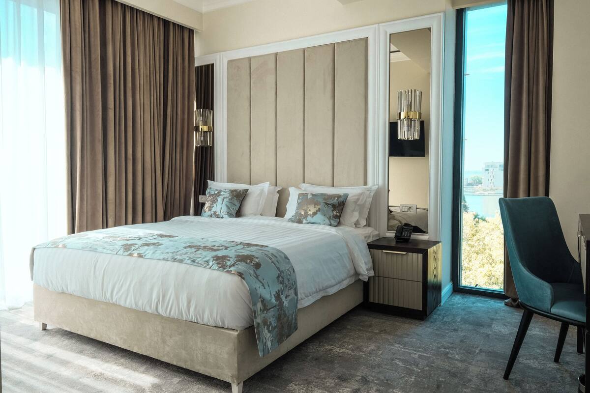 A modern bedroom with a large bed, a nightstand, a chair, and a view of the outdoors.
