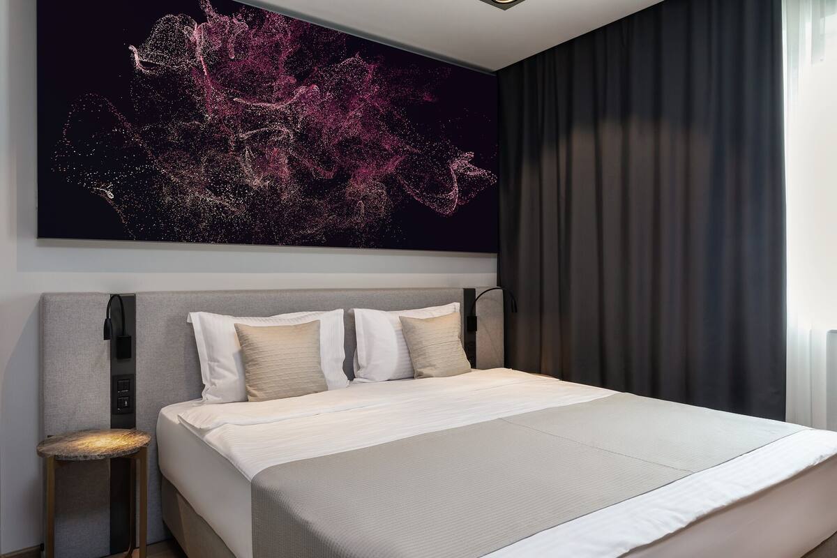 A modern hotel room with a large bed, a headboard with controls, a bedside table, and a large abstract painting on the wall.
