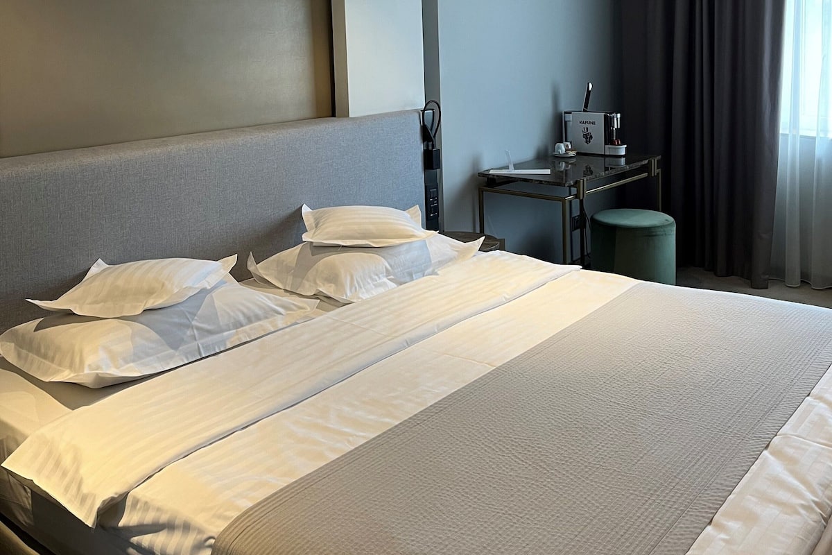 A hotel room with a large bed, a desk with a coffee machine, and a view of the outside through a window with curtains.