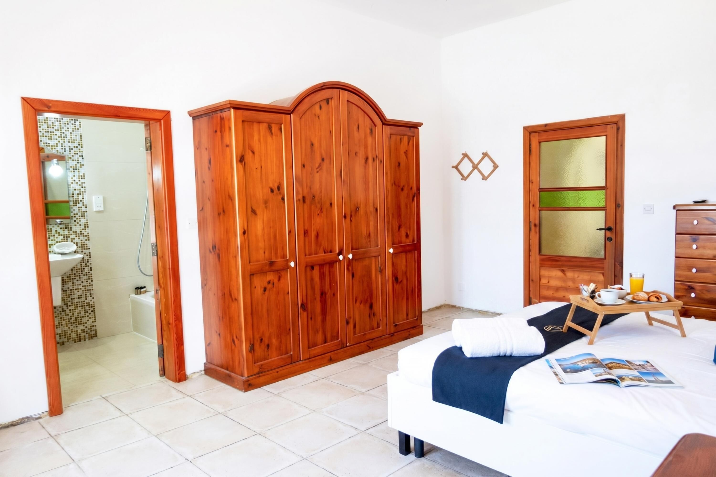 Een slaapkamer met een bed, een houten garderobe, een badkamer met een wastafel en een deur met een geëtste glazen panelen.